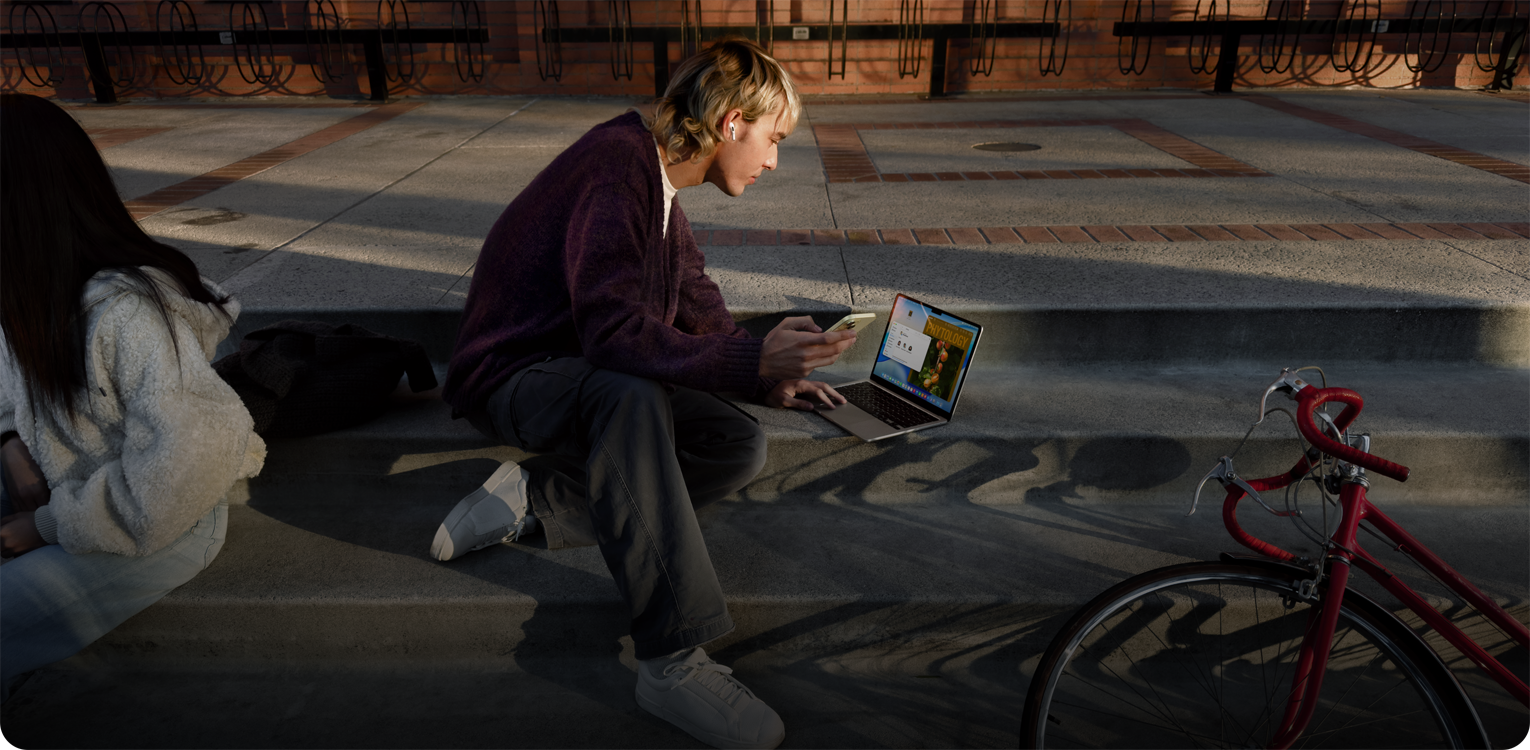 Person sitting on concrete steps outdoors, wearing an AirPod, holding an iPhone, and navigating on the trackpad of MacBook opened on the ground in front of them, red bicycle lying on floor nearby, another person sitting nearby