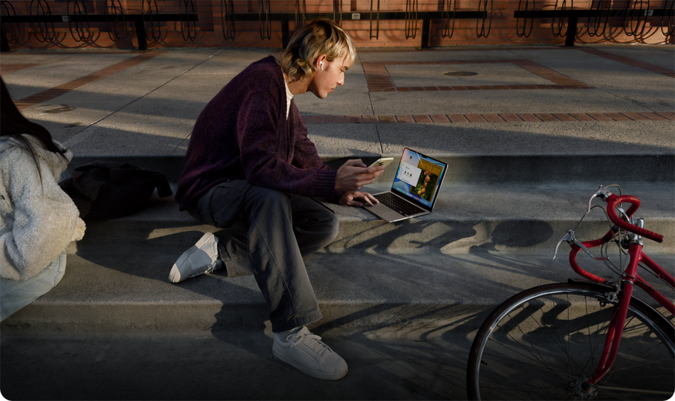 Person sitting on concrete steps outdoors, wearing an AirPod, holding an iPhone, and navigating on the trackpad of MacBook opened on the ground in front of them, red bicycle lying on floor nearby, another person sitting nearby